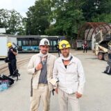 Marcel Grigull und Mirko Drotschmann posieren für ein Bild in einem Bergbauoutfit. Im Hintergrund ist ein Kamerateam zu erkennen und das Gelände des Trainingbergwerks Recklinghausen.