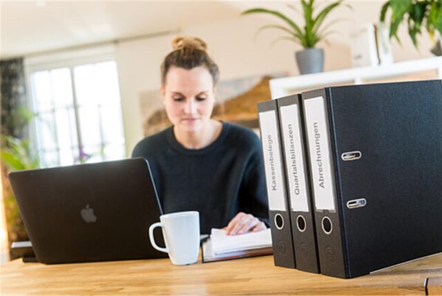 Eine Frau sitzt am Tisch und macht ihre Steuererklärung. Vor ihr stehen ein Laptop und Aktenordner.