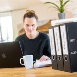 Eine Frau sitzt am Tisch und macht ihre Steuererklärung. Vor ihr stehen ein Laptop und Aktenordner.