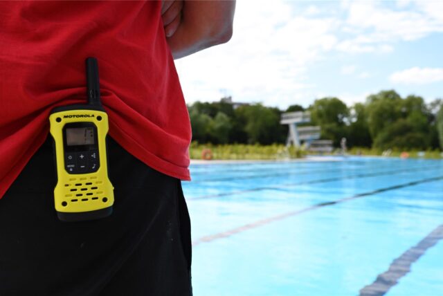 Der Rücken eines Rettungsschwimmers. Er trägt ein Funkgerät am Hosenbund. Im Hintergrund ist ein Freibad zu erkennen.