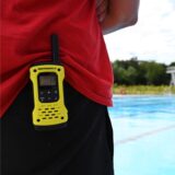 Der Rücken eines Rettungsschwimmers. Er trägt ein Funkgerät am Hosenbund. Im Hintergrund ist ein Freibad zu erkennen.