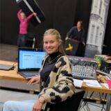 Maja Kost sitzt als Regieassistentin mit Laptop und Technikpult im Probenraum des WLT, im Hintergrund arbeiten zwei Personen an Bühnenelementen.