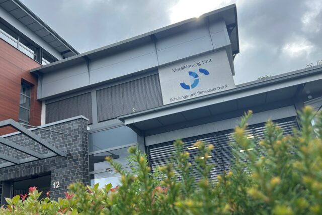 Das Bild zeigt die Fassade eines Gebäudes, auf dem das Schild „Metall-Innung Vest, Schulungs- und Servicezentrum“ angebracht ist. Im Vordergrund ist eine grüne Hecke zu sehen, die einen Teil des Gebäudes verdeckt.