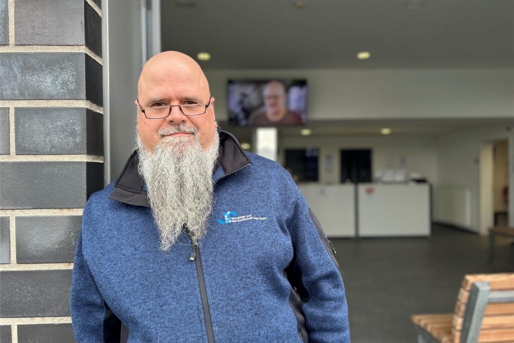 Ein Mann mit Brille und Bart steht vor einem Gebäude. Er trägt eine blaue Jacke mit dem Logo 