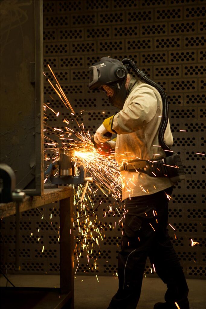 Ein Arbeiter schleift ein Metallstück, das in einem Schraubstock befestigt ist. Funken sprühen, während er Schutzkleidung und einen Helm trägt.