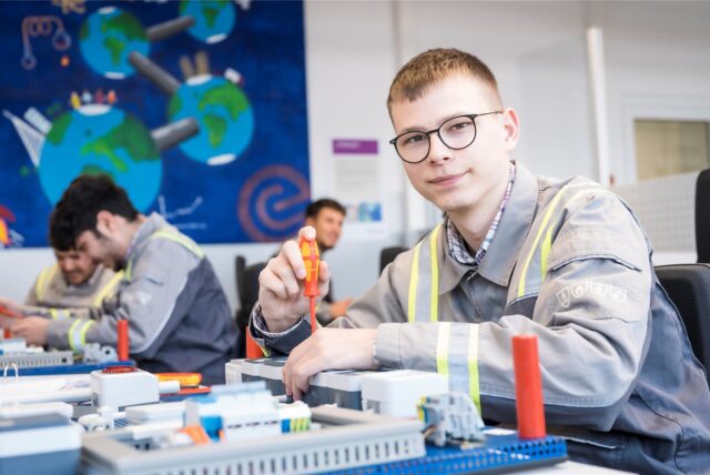Ein junger Mann mit Brille und Arbeitskleidung sitzt an einem Tisch und montiert mit einem Schraubendreher ein Modell. Im Hintergrund sind weitere Personen und eine Wandbemalung mit Erdkugeln zu sehen.