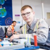 Ein junger Mann mit Brille und Arbeitskleidung sitzt an einem Tisch und montiert mit einem Schraubendreher ein Modell. Im Hintergrund sind weitere Personen und eine Wandbemalung mit Erdkugeln zu sehen.