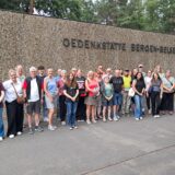 Gruppe von Teilnehmenden der „Tour de Demokratie“ steht vor der Wand mit dem Schriftzug „Gedenkstätte Bergen-Belsen“.