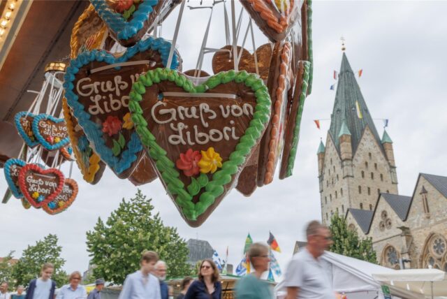 Lebkuchenherzen mit der Aufschrift „Gruß von Libori“ hängen an einem Stand der Libori-Kirmes in Paderborn. Im Hintergrund sind Besucher und der Dom zu sehen.