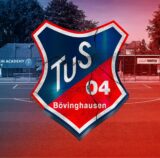 Im Bild ist das Logo des Tus Bövinghausen zu sehen, im Hintergrund ist der Fußballplatz zu sehen.