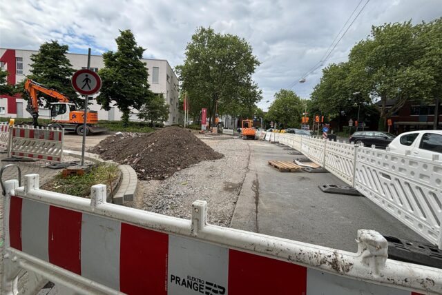 Straßenbaustelle mit weißen Absperrungen, einem Bagger und Erdaufschüttung, Zufahrt an einer Kreuzung teils gesperrt.