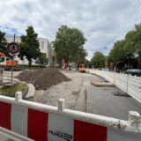 Straßenbaustelle mit weißen Absperrungen, einem Bagger und Erdaufschüttung, Zufahrt an einer Kreuzung teils gesperrt.
