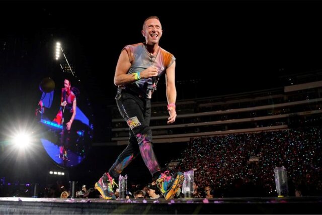 Chris Martin steht mit einem Mikrofon auf eine Bühne.