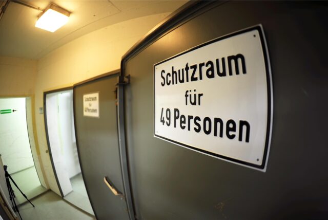 Man sieht ein Schild auf einer großen Tür eines Schutzraums. Darauf steht: „Schutzraum für 49 Personen“.