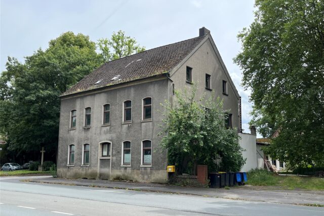 Ansicht eines zweistöckigen Wohnhauses mit grauer Fassade, das zwangsversteigert wird. Vor dem Haus befinden sich mehrere Mülltonnen und ein gelber Briefkasten.