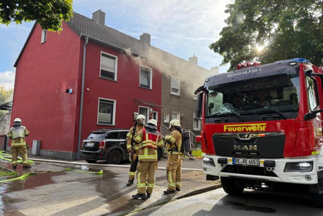 Feuerwehrleute besprechen sich neben einem Löschfahrzeug vor einem Wohnhaus in Recklinghausen stehend. Aus dem Haus an der Heinrichstraße dringt noch Rauch.